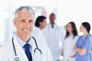 This is a picture of a male Doctor smiling. There is a blurred background of two female Nurses and two male Physicians talking.